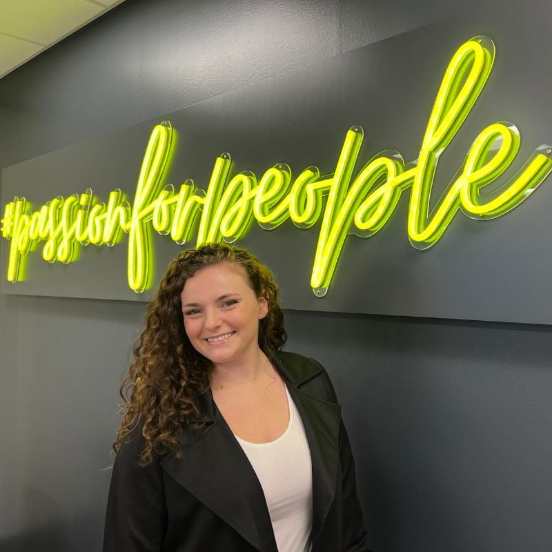 Brunel Account Coordinator Katie Rossmann smiles and poses in front of a neon yellow sign that reads #passionforpeople