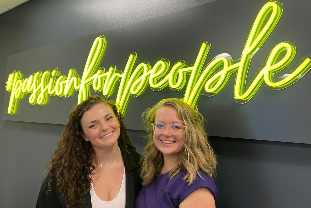 New Brunel Account Coordinators Katie Rossmann (left) and Abbi DeBeck (right) pose and smile together in front of neon yellow sign that reads "#passionforpeople"