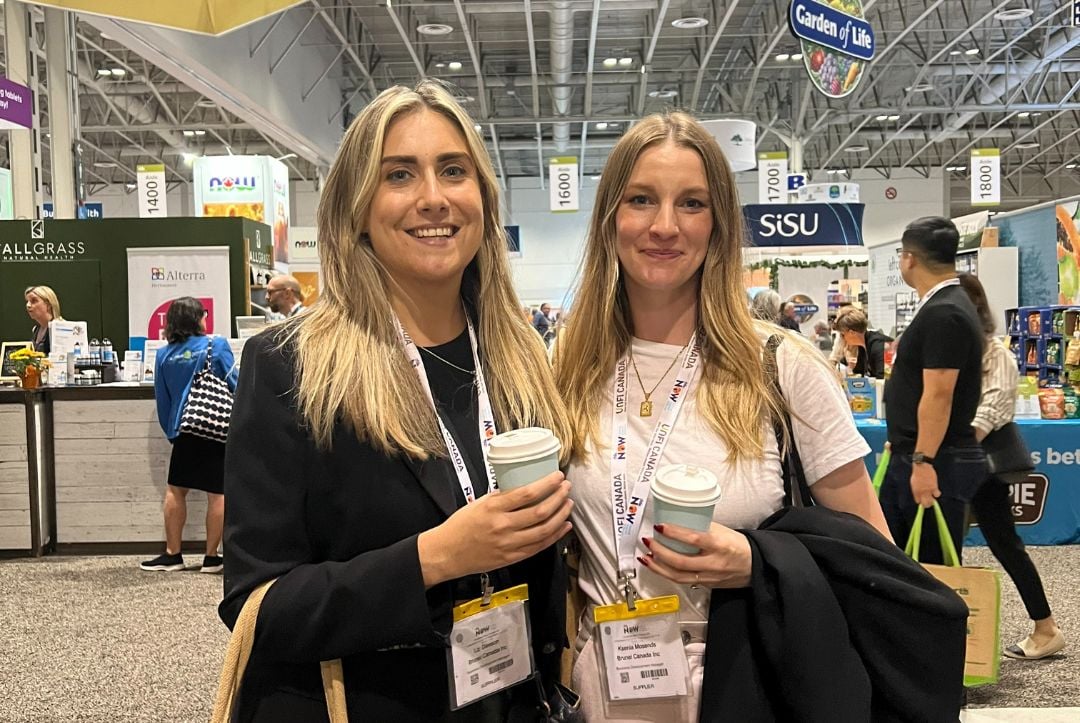 Women pose next to each other holding coffee mugs at a trade show