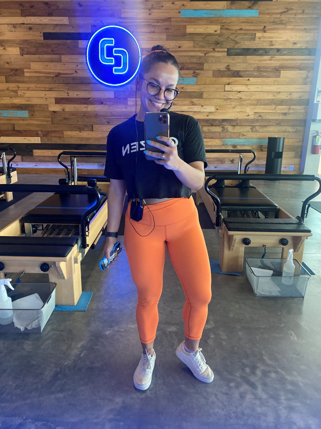 A woman in black t-shirt and orange leggings takes a selfie at a pilates studio