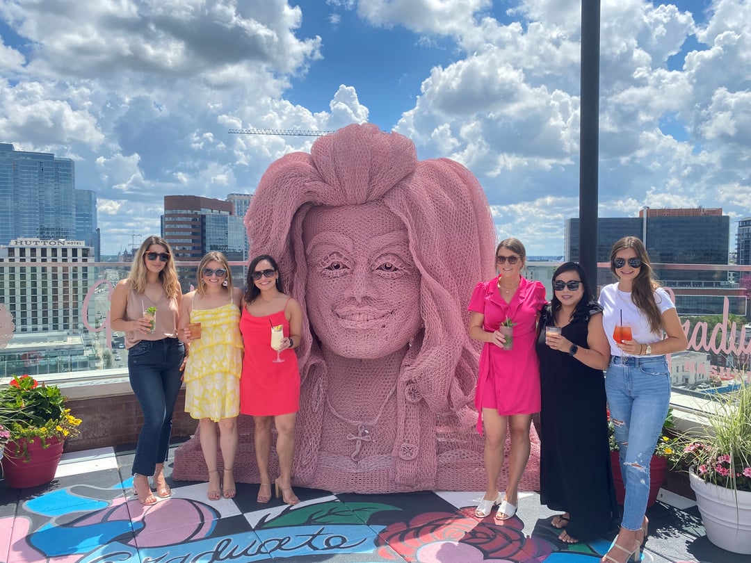 6 women stand next to pink art piece of woman's face, each holding drinks in their hands.