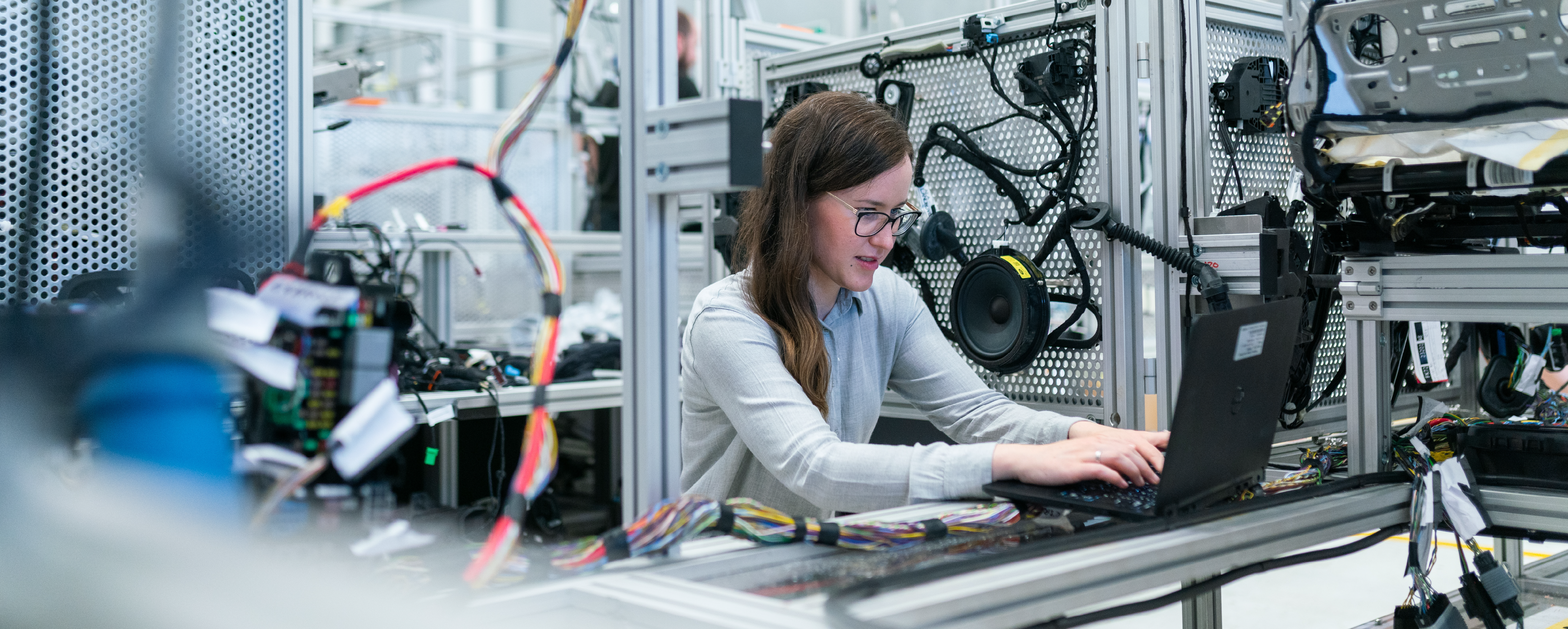 A Visionary at Work: Engineering a Sustainable Future A female engineer wearing glasses sits at a high-tech workstation, using a laptop to program or monitor complex electrical wiring and industrial components in a modern laboratory.