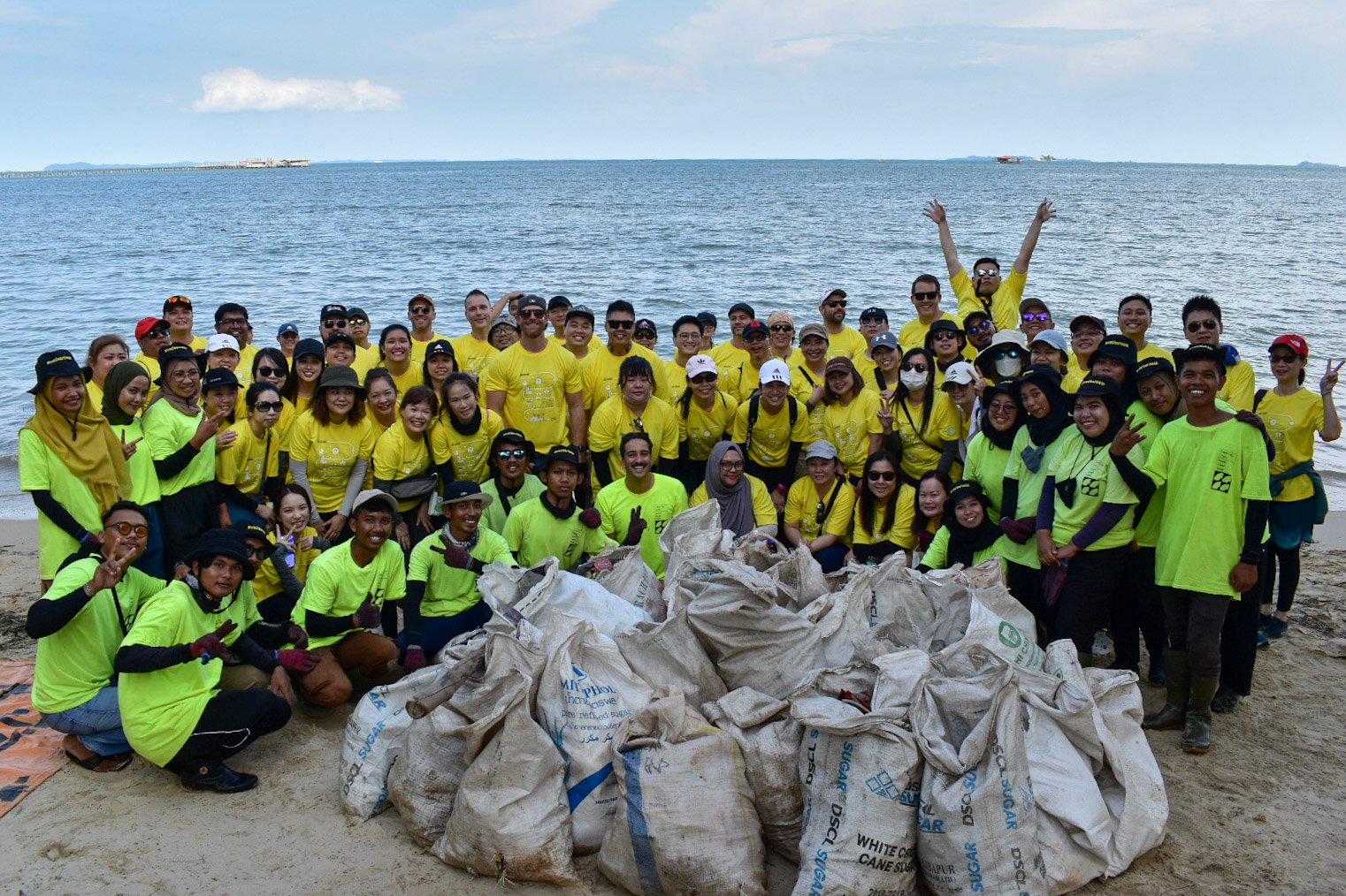 Beach Cleanup Volunteer Group Group of volunteers in yellow and green shirts on a beach posing with collected trash bags after a cleanup event.