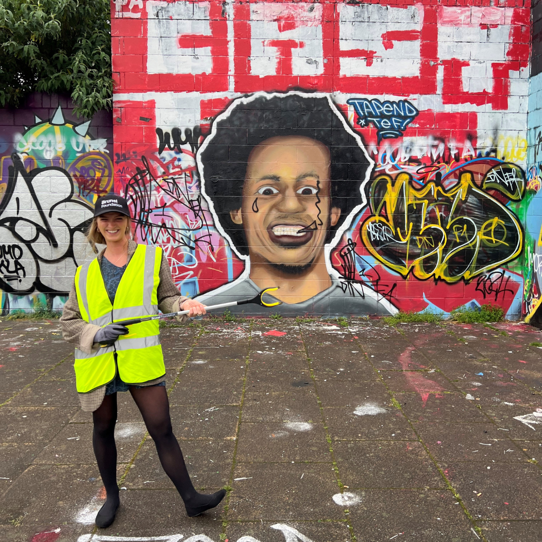 Brunel Foundation Trash ’n Trace Clean-up Brunel volunteer wearing a yellow safety vest holding a litter picker in front of a colorful graffiti wall during a Trash ’n Trace Clean-up activity.