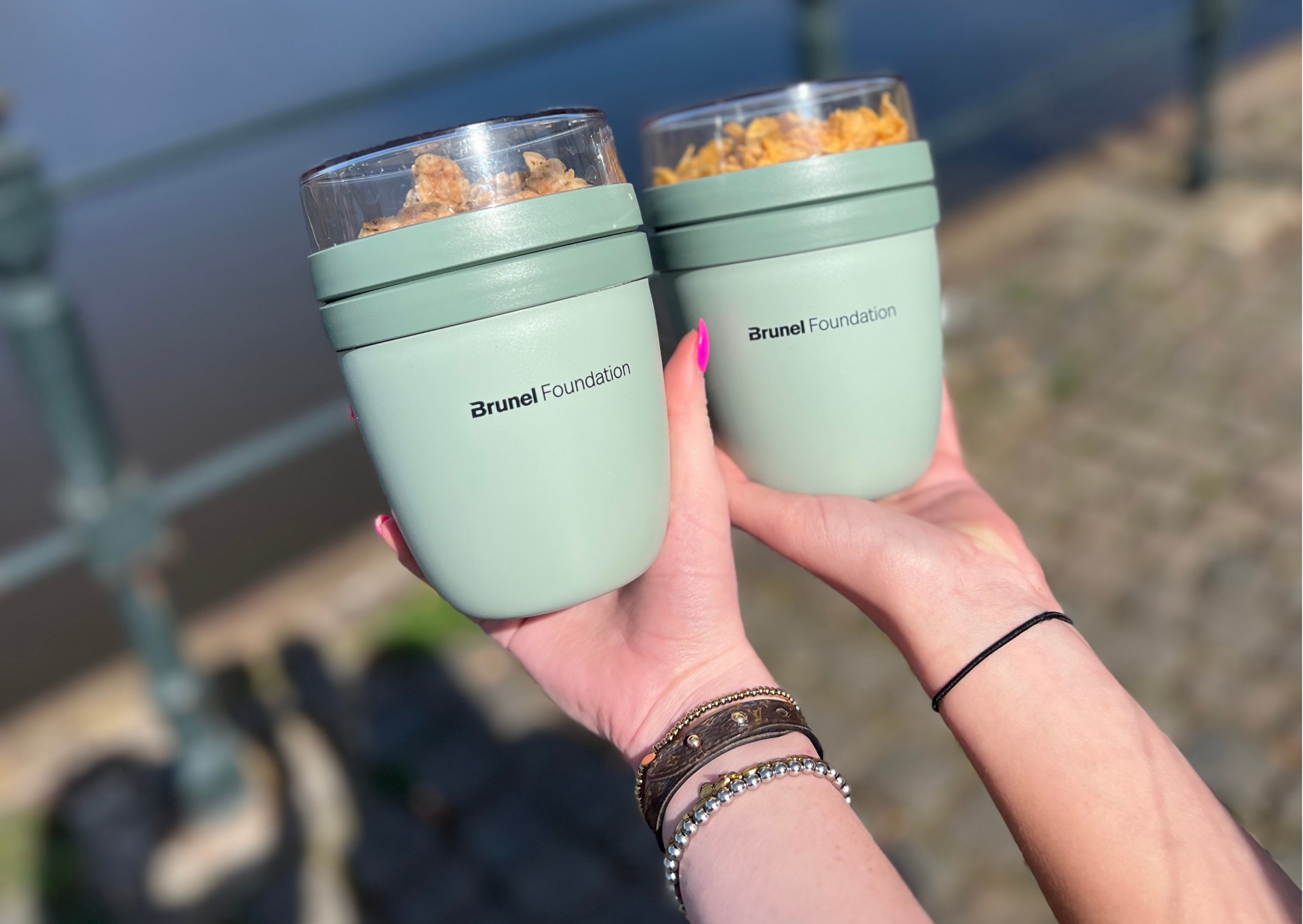 Brunel Foundation Reusable Cups Two reusable Brunel Foundation cups filled with snacks held outdoors near a waterway.