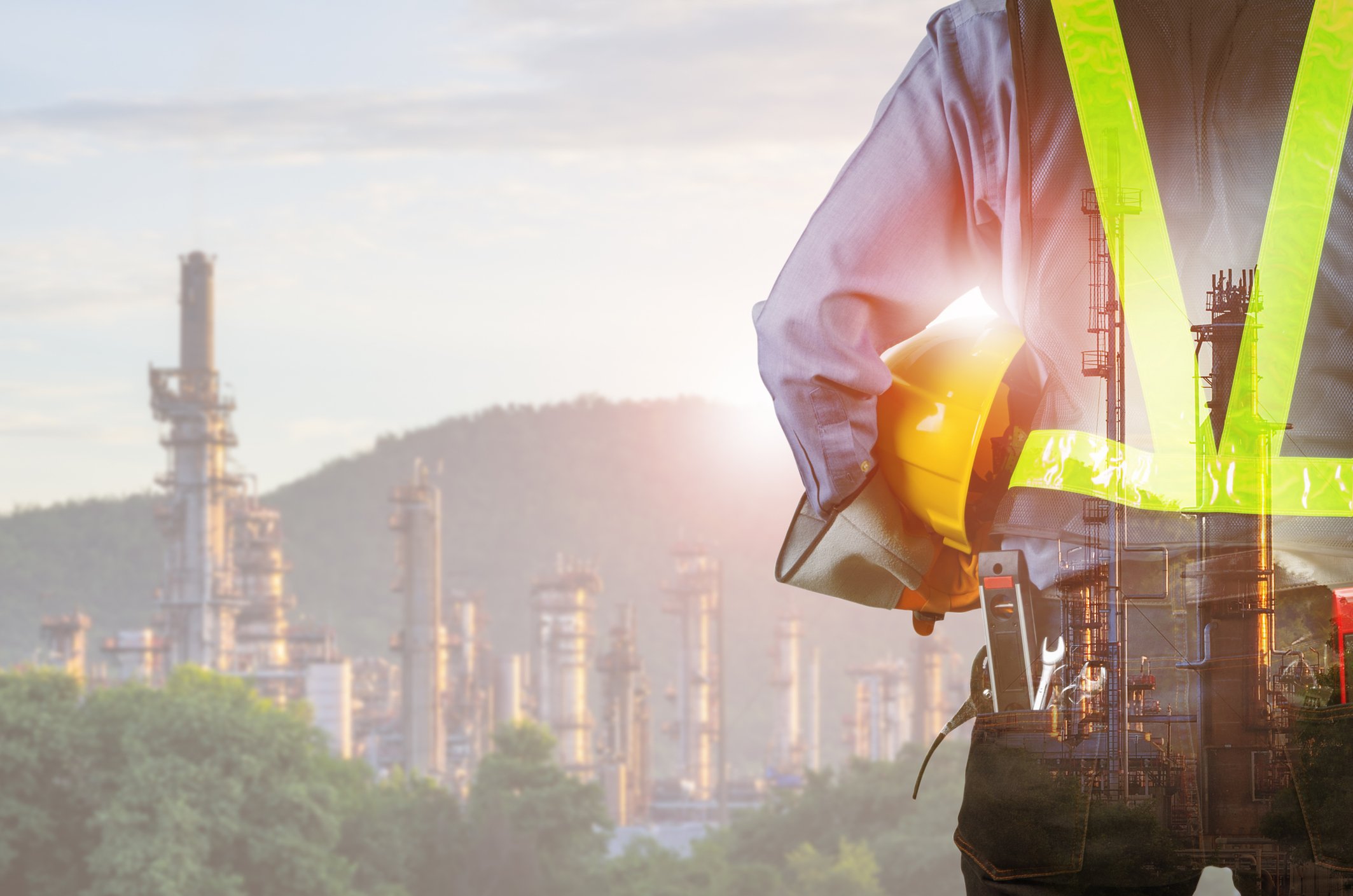 Worker in high‑visibility vest holding a hard hat overlooking refinery towers at sunrise, safety culture in oil rig jobs