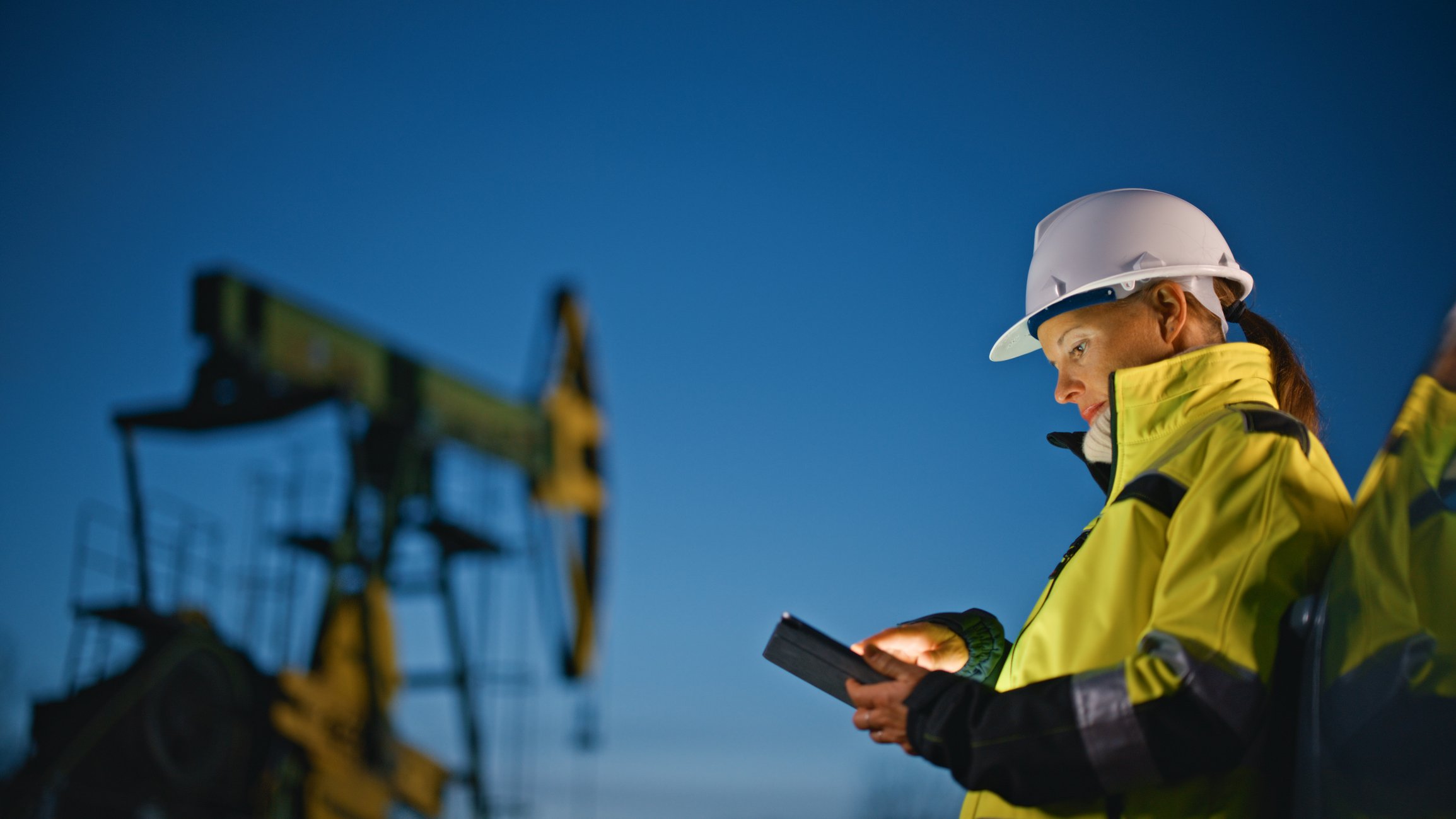 Oilfield technician using a tablet near a pumpjack at dusk, tech skills and data workflows in modern oil rig jobs