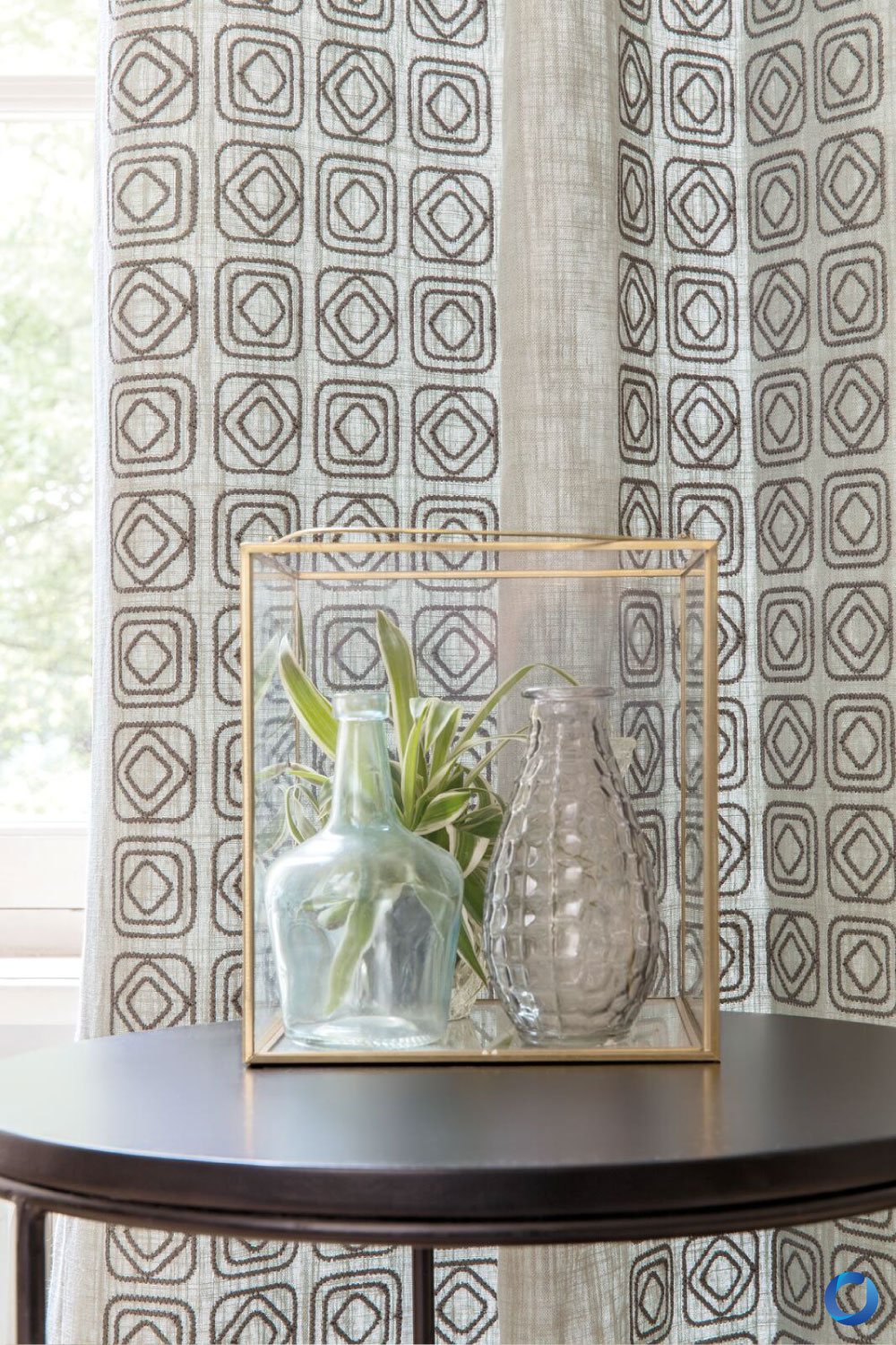 Close-up of a decorative table featuring glass vases, a plant, and a gold-framed accent piece. The backdrop showcases geometric-patterned curtains in earthy tones, creating a sophisticated and artistic display.