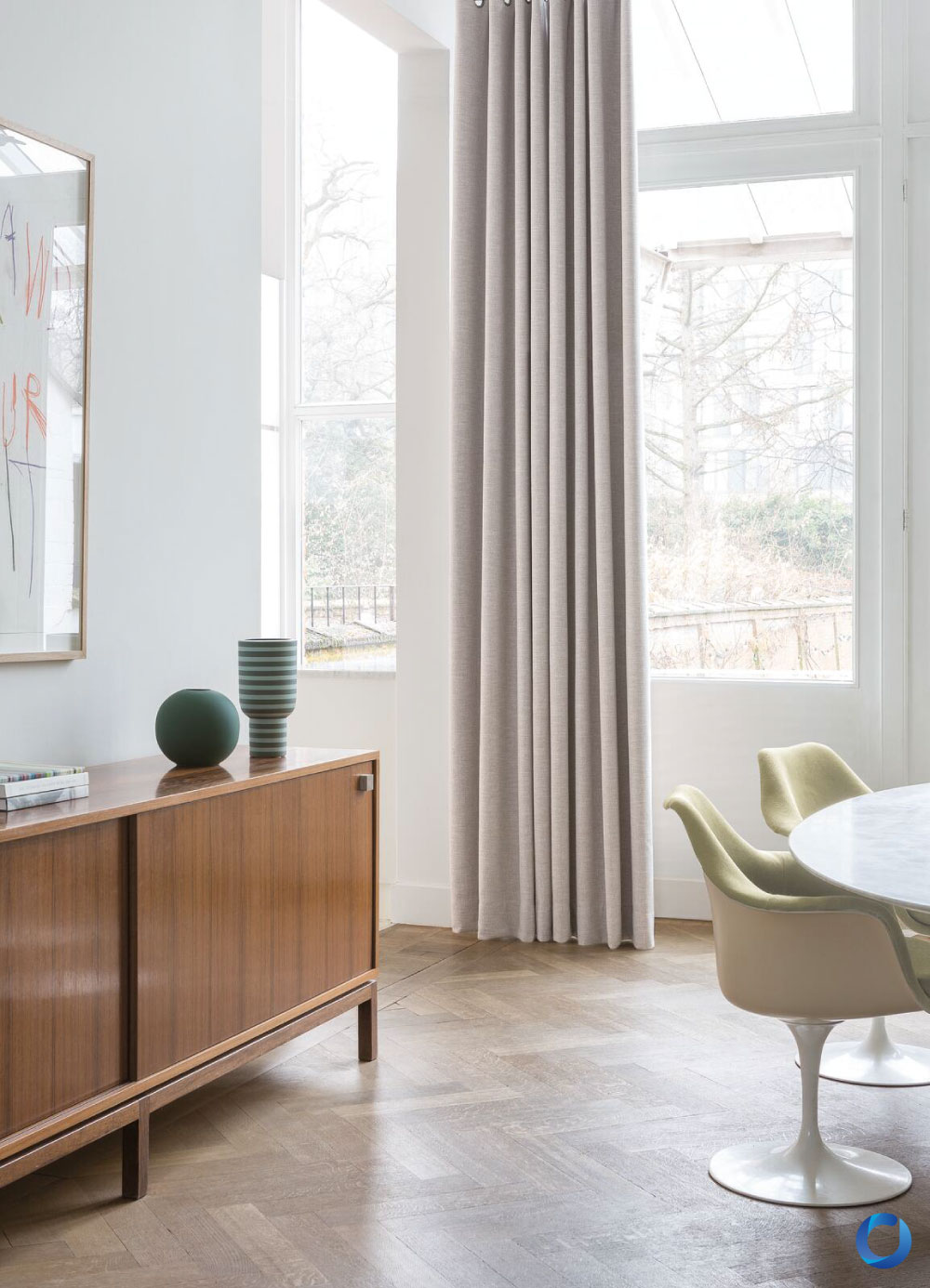 Minimalist dining space with light wood flooring, beige floor-length curtains, a mid-century wooden sideboard, and modern decor. The room features natural light, a marble-top table, and stylish green chairs.