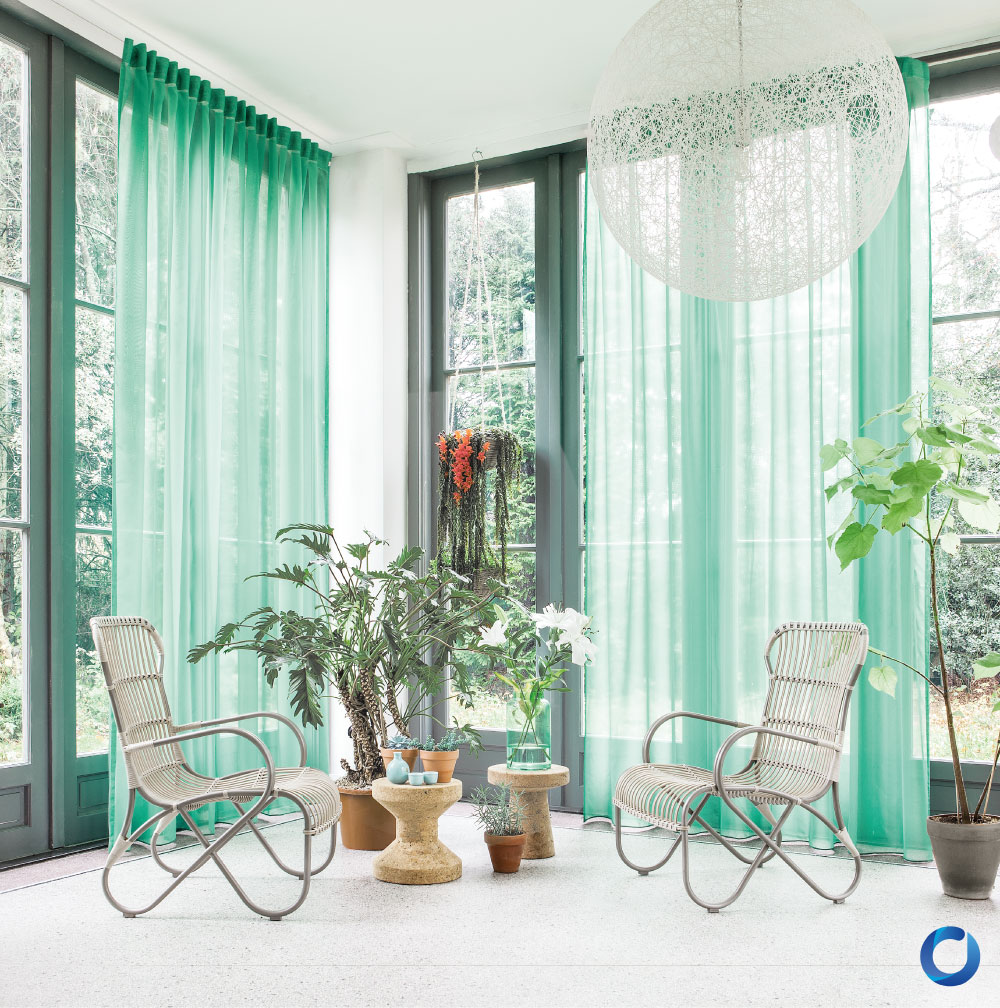 Bright sunroom featuring floor-to-ceiling green sheer curtains, rattan chairs, and potted plants. The space is illuminated by natural light, creating a fresh, serene ambiance with natural decor elements.