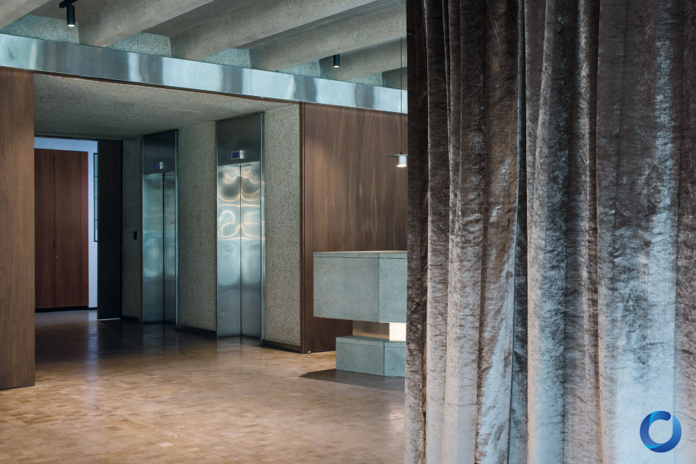Contemporary interior with metallic elevators, concrete accents, wood paneling, and plush velvet curtains. The space features a minimalist design with a blend of industrial and luxurious elements.