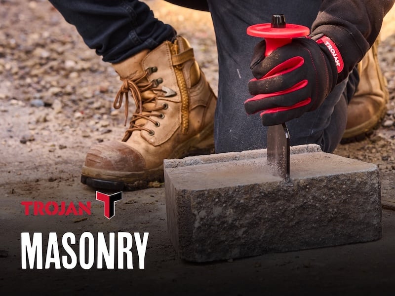 Person in jeans and boots using a brick bolster to cut stone with the text "Masonry" in the foreground