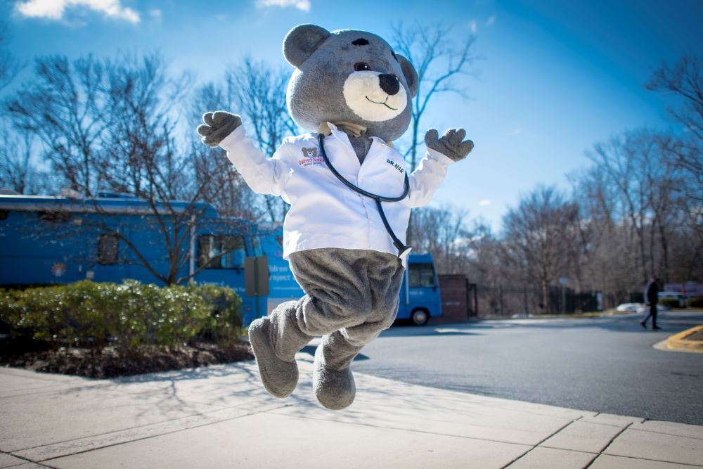 Children's National Hospital mascot Dr. Bear jumps in the air against a bright blue sky background