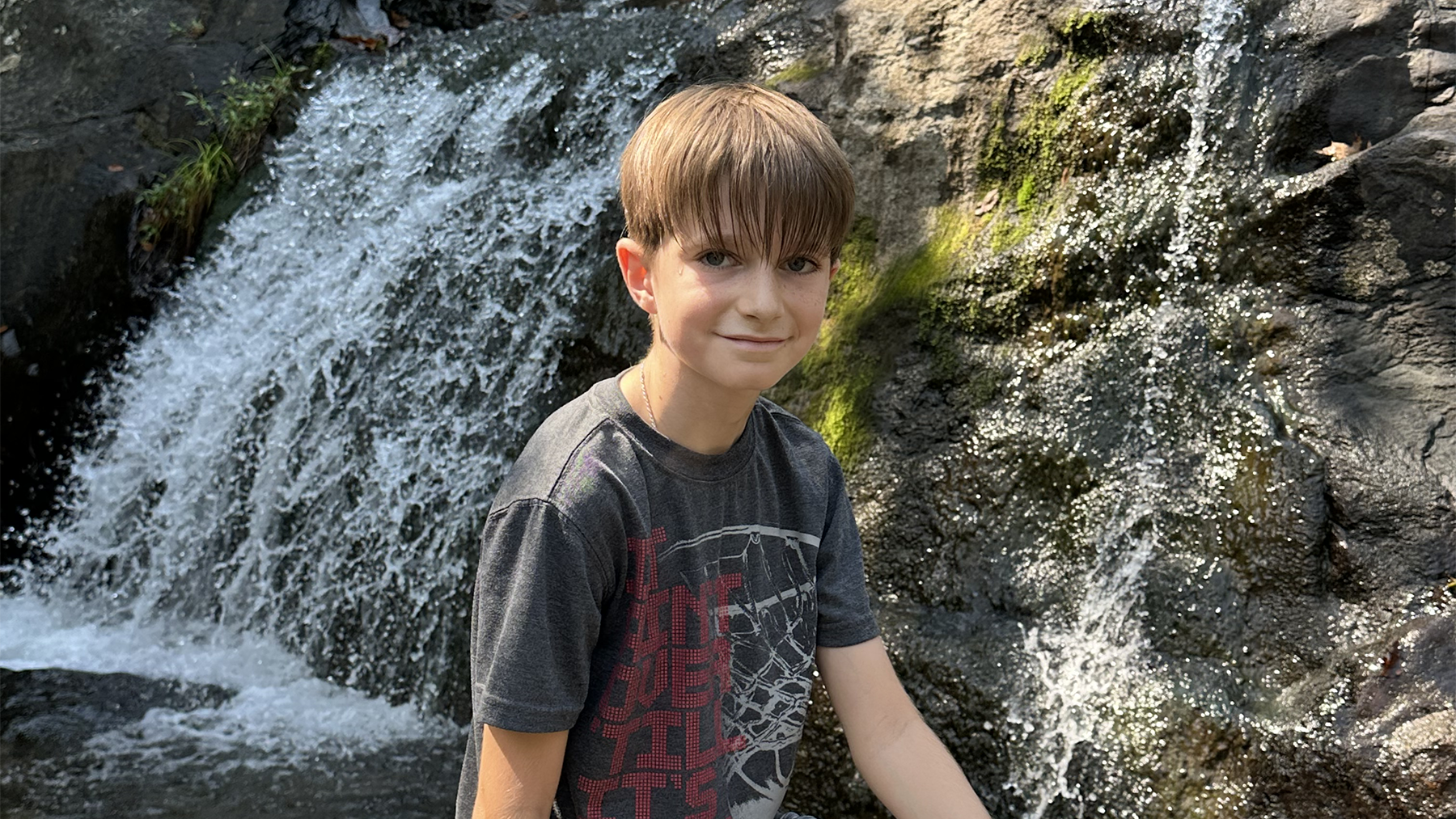 Boy wearing a t-shirt sits in front of a waterfall