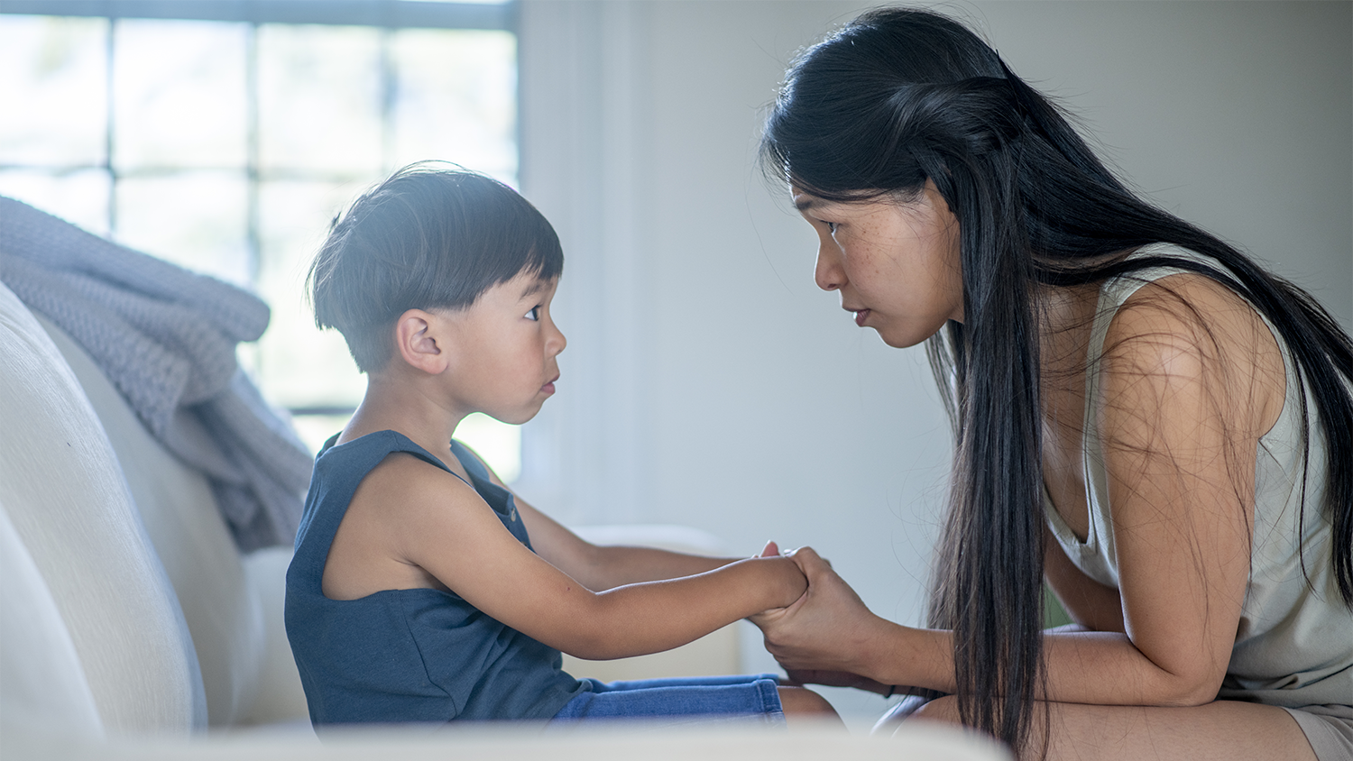 A young brown-haired boy in a blue shirt holds his mother's hands and looks at her.