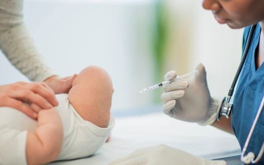 A nurse in blue scrubs gives a baby a vaccination shot in his thigh.