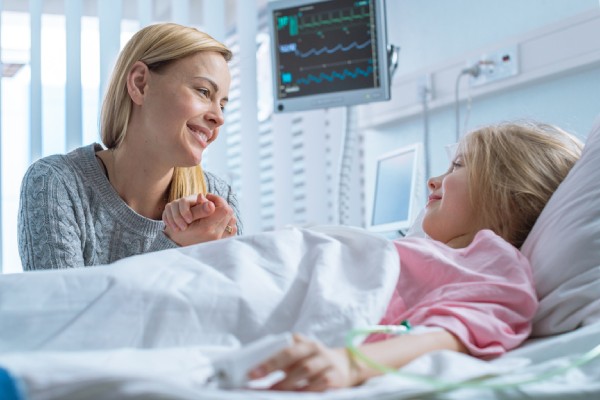 Visiting mother holding hand of young smiling daughter laying in a hospital bed