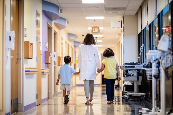 A boy and girl hold a provider's hands and walk down a hallway at the main hospital.