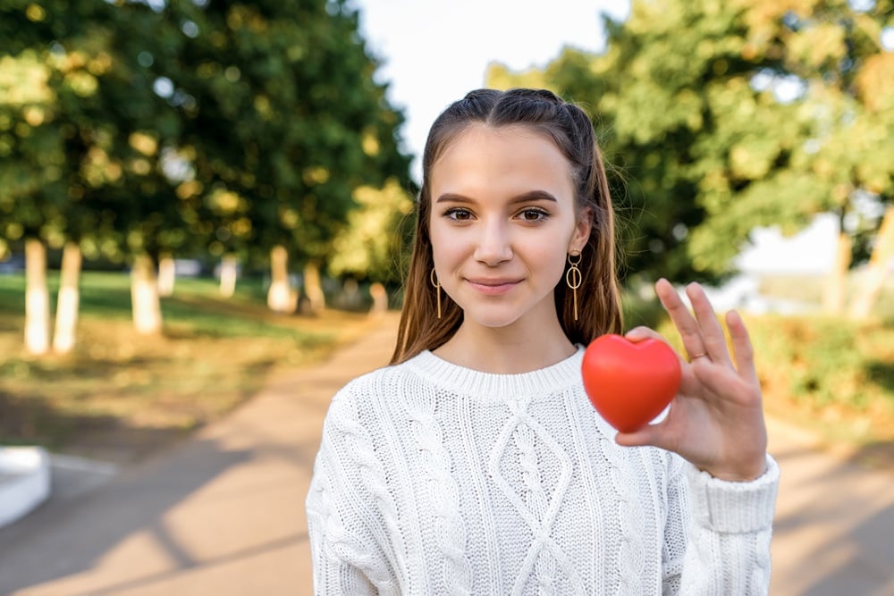 A teen girl standing on a tree-lined street holding a small red heart