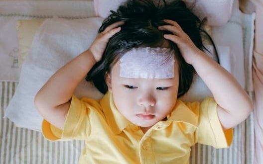 A young girl in a yellow shirt lays on a pillow with her hands on her head with a headache.