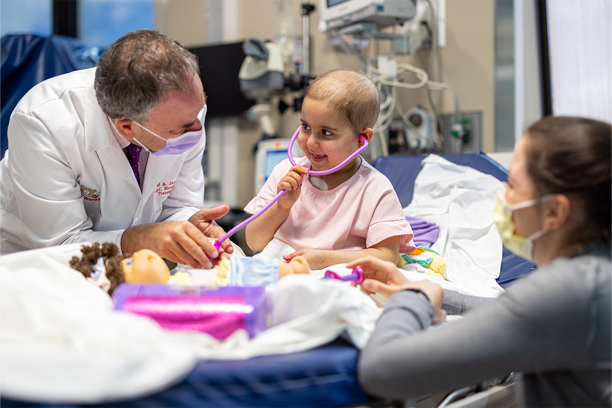 young girl with cancer in hospital bed plays with a doll and her two doctors