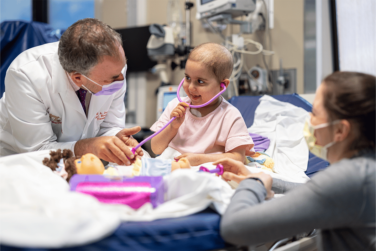 young girl with cancer in hospital bed plays with a doll and her two doctors