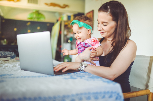 A smiling baby girl sits on her mother's lap as mom works on a laptop at her kitchen table