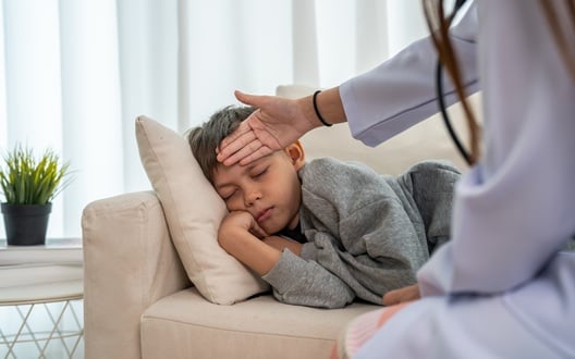 A child rests on a couch while their parent holds a hand to their forehead to check for a fever