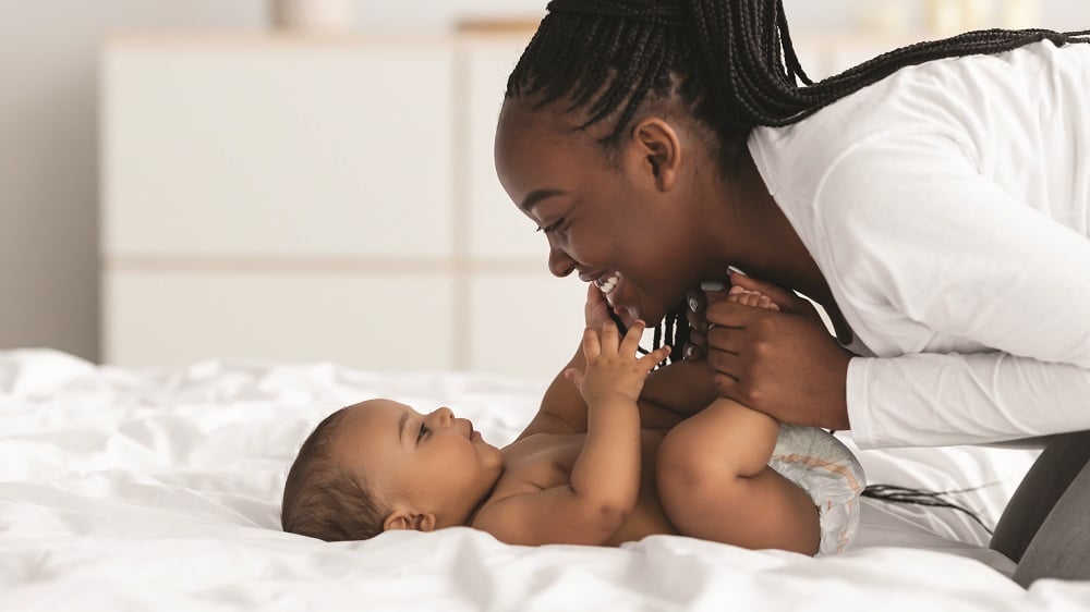 A young mother leans over her baby laying on a bed and smiles while holding her baby's feet.