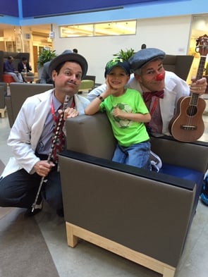 Brayden smiling while sitting next to two doctor clowns holding musical instruments at Children's National Hospital