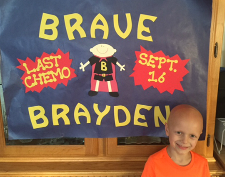 Brayden with a bald head standing in front of a sign that says "Brave Brayden" and lists the date of his last chemotherapy appointment