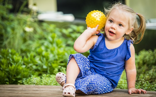 A toddler girl holds a yellow ball while sitting on a bench in the healing garden at Children's National Hospital.