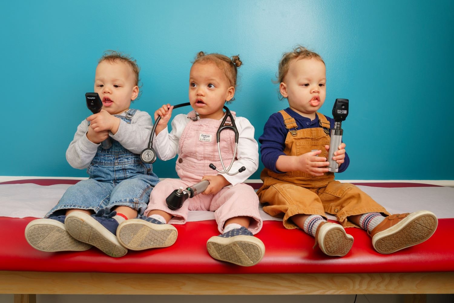 triplets sit on exam table playing with doctor's equipment