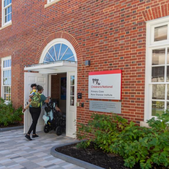 Photograph of a woman and two children entering the Children's National Shepherd Park Primary Care office main entrance in Washington, D.C. The woman is carrying one child and the other child is being pushed in a wheelchair.