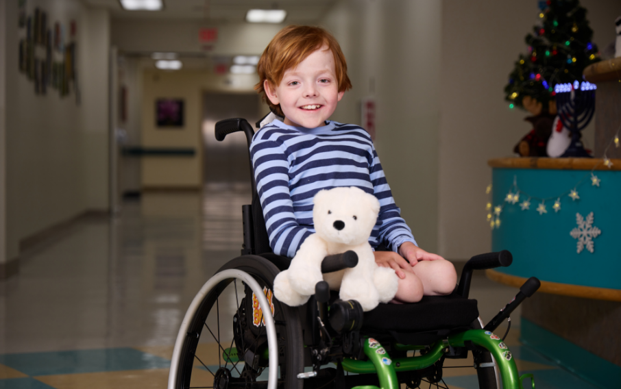 A young patient in a wheelchair smiles at the camera accompanied by a polar bear plush toy