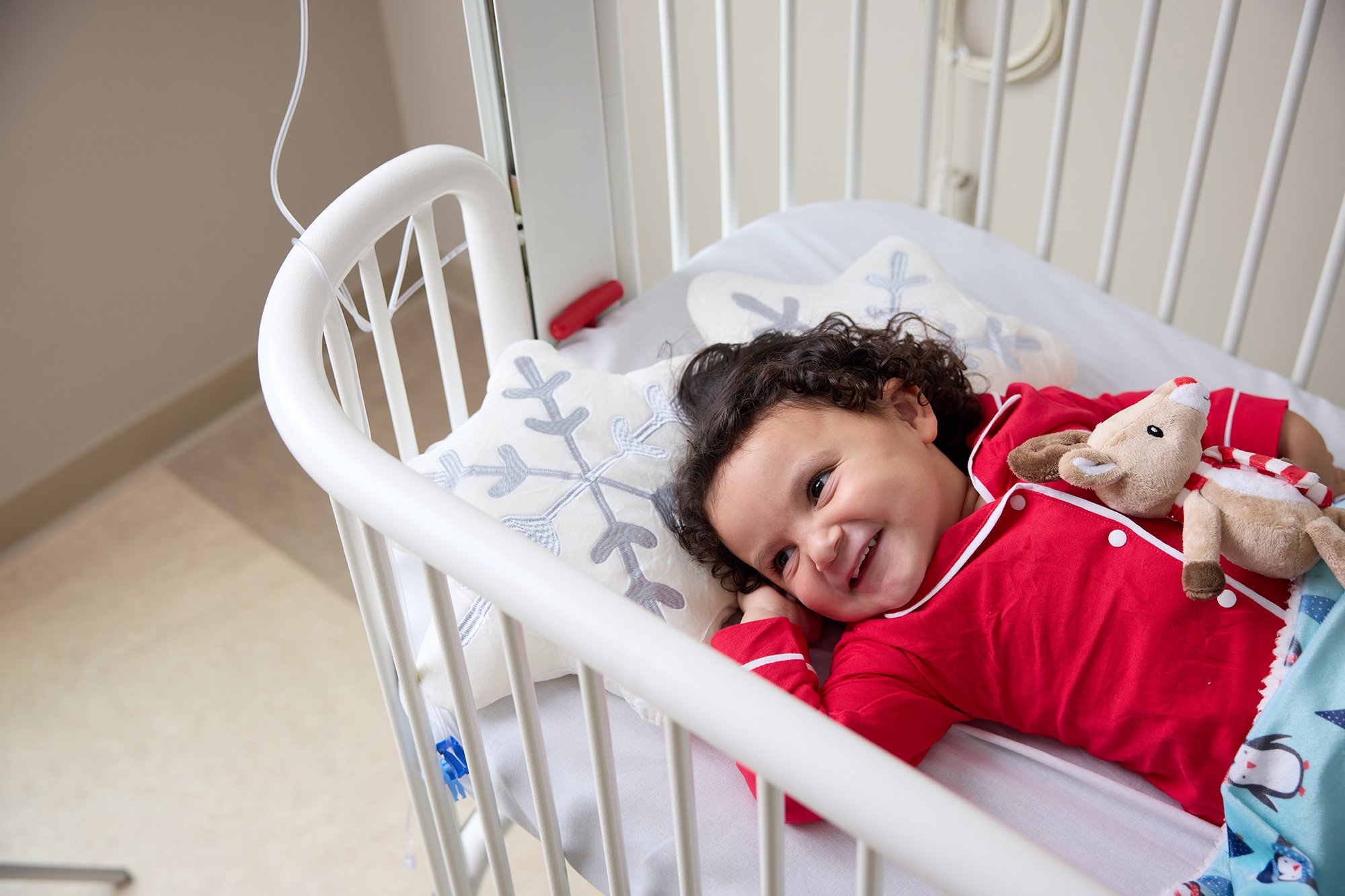 CNH patient EmmyLou, age 3, laying in a crib bed while hugging a reindeer stuffed animal and wearing red pajamas.