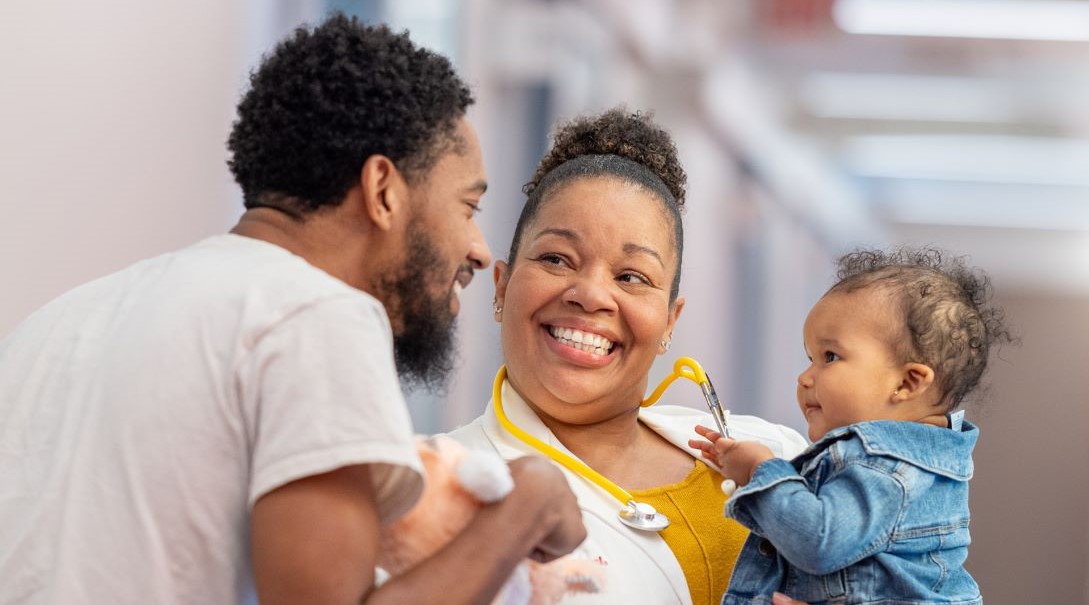 An adult female doctor smiles while holding an infant who is playing with her stethoscope.
