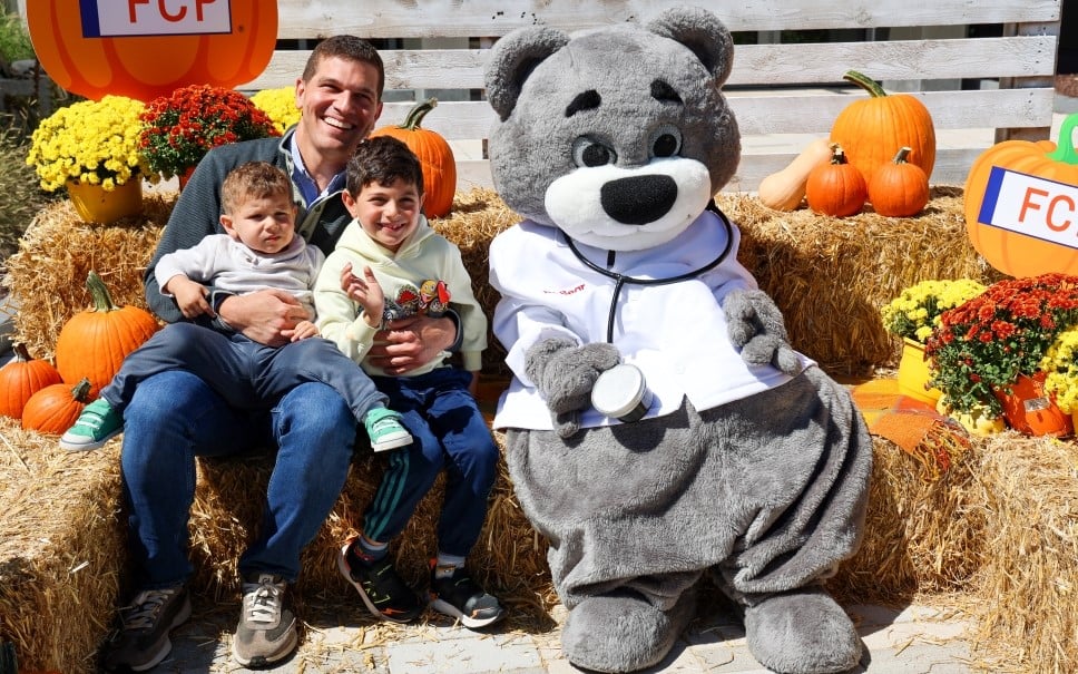 Children's Hospital mascot, Dr. Bear, sitting on hay bales with a father and two young children at the 2024 Champions for Children's National Fall Fest.