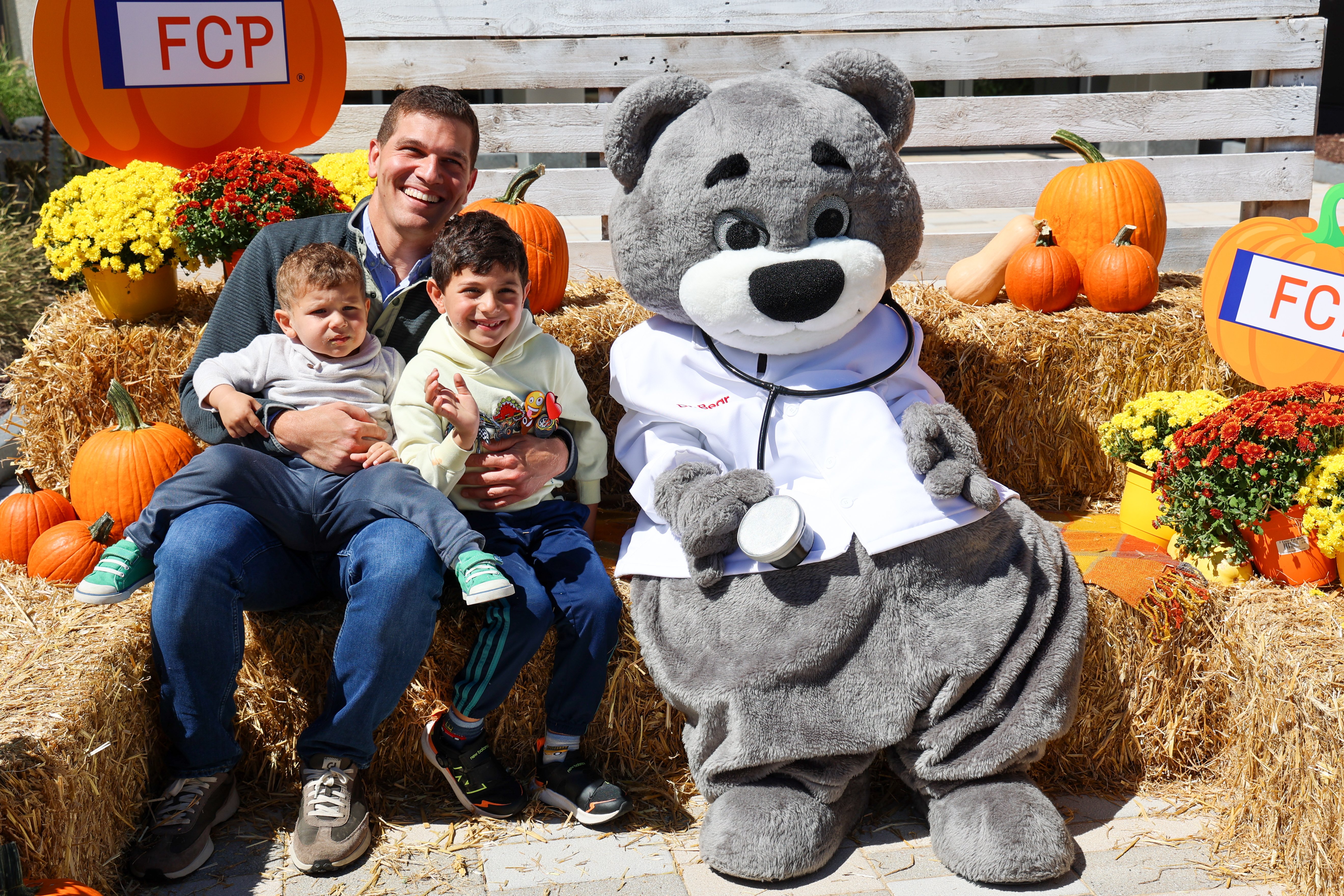 Children's Hospital mascot, Dr. Bear, sitting on hay bales with a father and two young children at the 2024 Champions for Children's National Fall Fest.