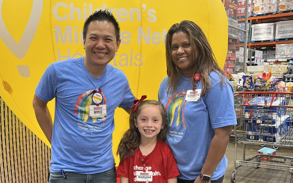 Two adults and a female child stand in front of a yellow Children's Miracle Network Hospitals display inside a Costco.