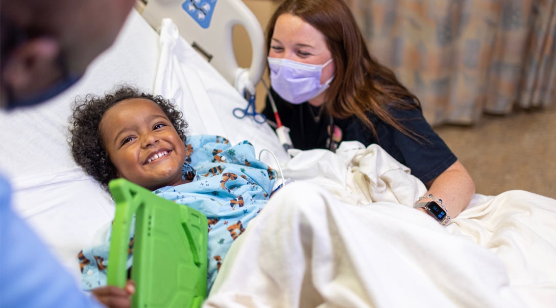 A Black pre-school aged patient smiles in their hospital bed while a White female caregiver looks on and smiles.