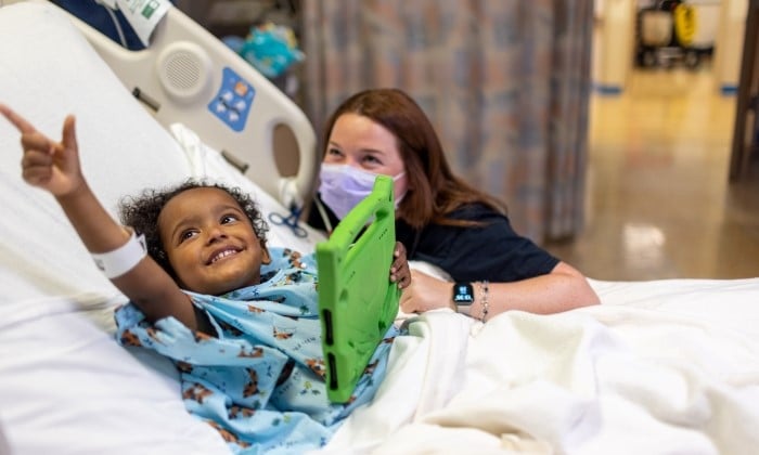 A Black pre-school aged patient smiles in their hospital bed while a White female caregiver looks on and smiles.