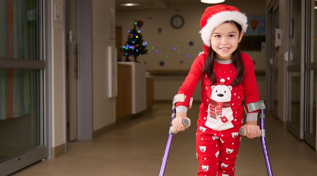 Aila, a young female patient utilizing crutches, smiles at the camera while wearing red winter pajamas and a Santa Claus hat