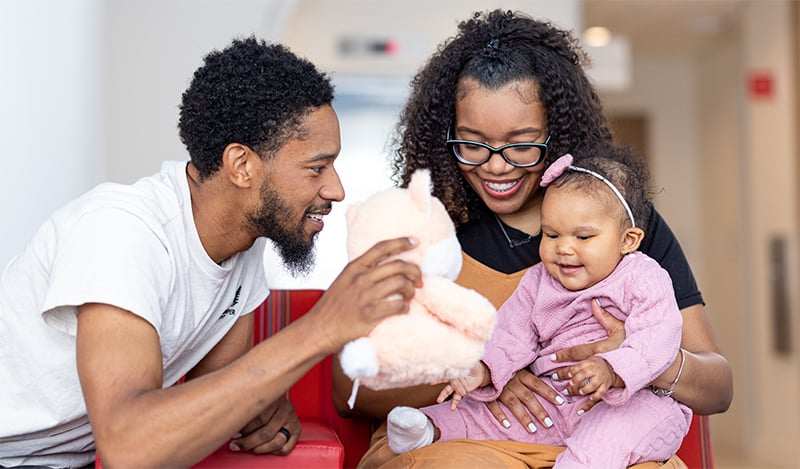 Woman holding smiling infant girl while adjacent man entertains the baby with a plush doll