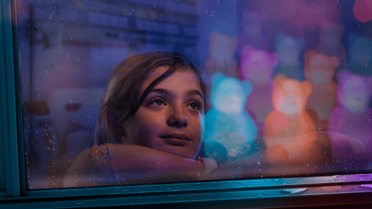 An elementary school aged female patient looks longingly out a window with the glow of Light Up Dr. Bears in the reflection.