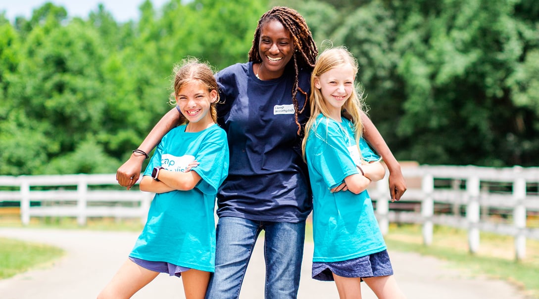 An adult female camp counselor laughs while posing with two smiling female campers.
