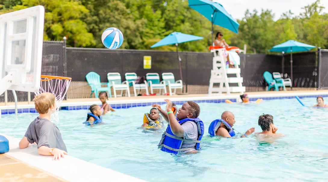 Campers of varying ages and abilities swim and shoot baskets in a pool while a lifeguard observes from a distance.