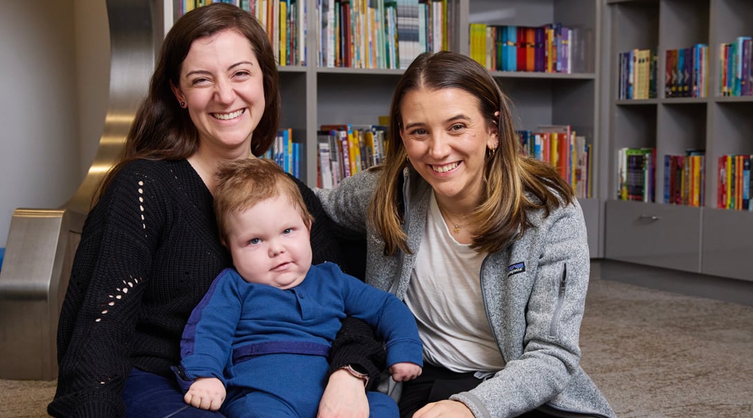 An infant patient, Jack, smiles while sitting with his mother, Rachel, and Child Life Specialist Lanie Berk.