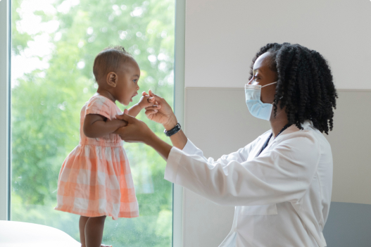Provider wearing a facemask holds the hands of a little girl who is standing on top of the exam table.