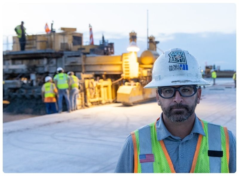 A man with a McNeil Brothers helmet standing in front of group of construction workers and a paving machine